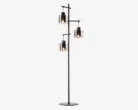 Høj standerlampe med mat sort metalstel og tre forskudte arme, der hver bærer en cylindrisk glasskærm. Glasskærmene er klare med en mat nederste halvdel, og hver skærm indeholder en synlig glødepære, hvilket giver et moderne, industrielt udtryk. Lampen står på en rund, flad fod for stabilitet.