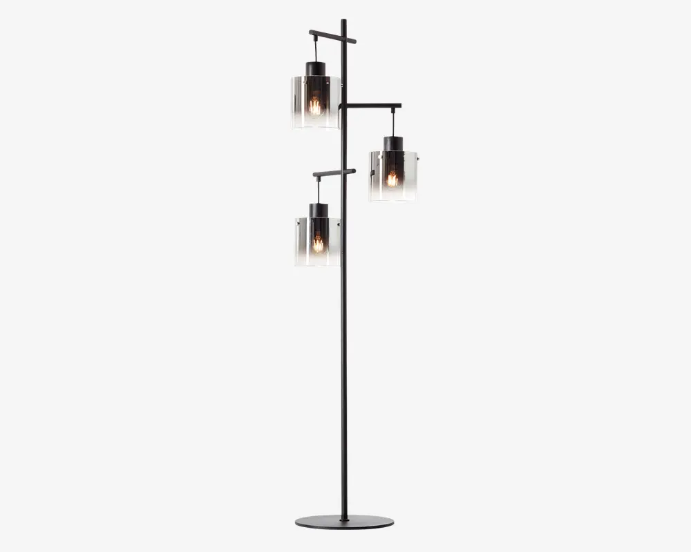Høj standerlampe med mat sort metalstel og tre forskudte arme, der hver bærer en cylindrisk glasskærm. Glasskærmene er klare med en mat nederste halvdel, og hver skærm indeholder en synlig glødepære, hvilket giver et moderne, industrielt udtryk. Lampen står på en rund, flad fod for stabilitet.