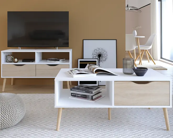 Rektangulært sofabord i moderne skandinavisk design med hvid ramme og front på skuffen i lyst træ. Bordet har skrå træben, en åben hylde til opbevaring og én skuffe med udskåret greb. På bordpladen står en glasvase, en lille skål og et åbent magasin, mens hylden nedenunder rummer flere stablede bøger. Udtrykket er minimalistisk og funktionelt.