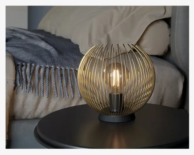 En kugleformet bordlampe med moderne design, bestående af tynde, jævnt fordelte metalstænger i guld, der danner et bur omkring en synlig pære. Lampens fod er sort og rund, hvilket giver en stabil base. Lampen udsender et varmt, hyggeligt lys og egner sig godt som natbordslampe eller stemningsbelysning.