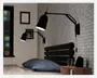 En moderne, sort vægmonteret lampe med justerbar arm og konisk skærm. Lampen er fastgjort til en hvid murstensvæg over en seng, hvilket gør det muligt at rette lyset efter behov. Designet er minimalistisk og funktionelt, velegnet som læselampe eller arbejdsbelysning ved sengen.