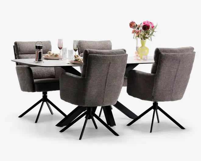 Et moderne spisesæt med et rektangulært bord med en glat, lys bordplade og sorte skrå ben. Sættet inkluderer fire polstrede stole i blødt gråt stof, hver med høj ryg, polstrede armlæn og sorte metalben med drejefunktion. Bordet er dækket med tallerkener, glas, bestik og en stempelkande, og pyntet med en vase med friske blomster, hvilket skaber en indbydende og stilfuld spiseatmosfære.