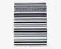 Rektangulært tekstil med vandrette striber i sort, hvid, grå og brune nuancer. Striberne varierer i bredde og har geometriske mønstre som prikker, diamanter og linjer. Kanterne er afsluttet med korte hvide frynser i begge ender.