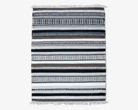 Rektangulært tekstil med vandrette striber i sort, hvid, grå og brune nuancer. Striberne varierer i bredde og har geometriske mønstre som prikker, diamanter og linjer. Kanterne er afsluttet med korte hvide frynser i begge ender.