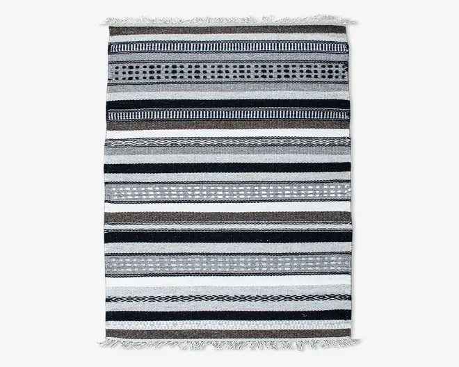 Rektangulært tekstil med vandrette striber i sort, hvid, grå og brune nuancer. Striberne varierer i bredde og har geometriske mønstre som prikker, diamanter og linjer. Kanterne er afsluttet med korte hvide frynser i begge ender.