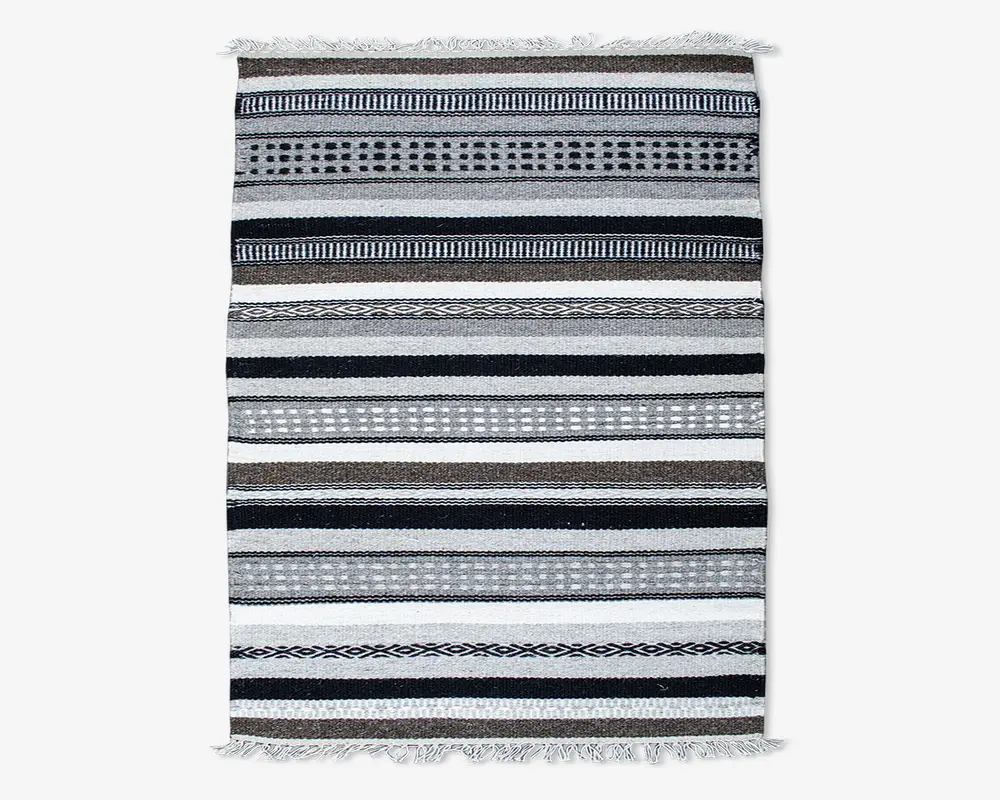 Rektangulært tekstil med vandrette striber i sort, hvid, grå og brune nuancer. Striberne varierer i bredde og har geometriske mønstre som prikker, diamanter og linjer. Kanterne er afsluttet med korte hvide frynser i begge ender.