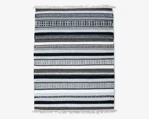 Rektangulært tekstil med vandrette striber i sort, hvid, grå og brune nuancer. Striberne varierer i bredde og har geometriske mønstre som prikker, diamanter og linjer. Kanterne er afsluttet med korte hvide frynser i begge ender.