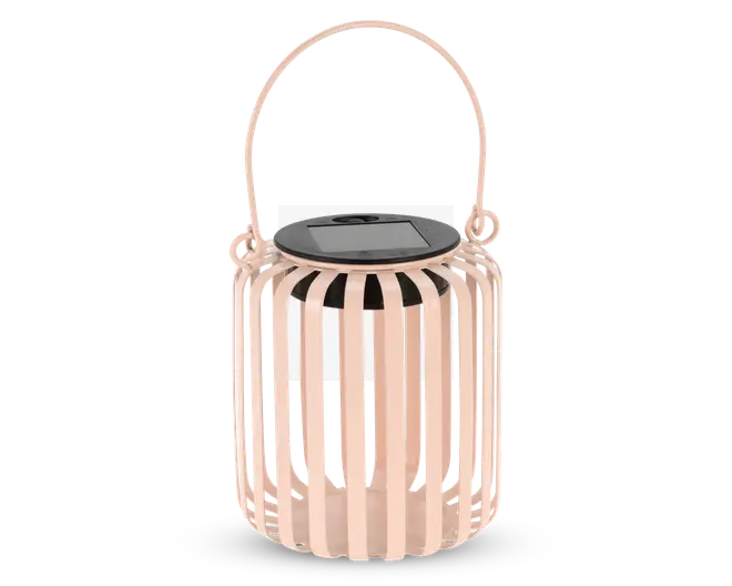 En dekorativ lanterne med en lyserød finish, der har lodrette lameller, som danner en cylindrisk form. Lanternen har et solidt, matchende håndtag øverst, så den nemt kan bæres eller hænges op. Øverst sidder et sort solpanel, hvilket viser, at lanternen er solcelledrevet. Det åbne lameldesign lader lyset skinne igennem og skaber et mønstret lys.
