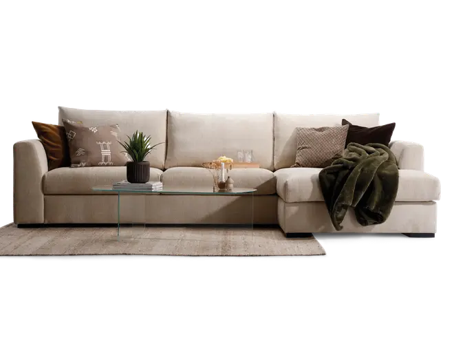 En moderne, lysebeige hjørnesofa med brede armlæn og bløde puder. Sofaen har en chaiselong på højre side og er pyntet med forskellige dekorative puder i neutrale og jordnære farver, herunder geometriske og teksturerede designs. Et mørkegrønt, blødt tæppe er lagt over chaiselongen. Foran sofaen står et slankt, rektangulært sofabord af glas med metalben, hvor der er placeret en potteplante, bøger, snacks og glas. Hele opstillingen står på et lyst, tekstureret tæppe, hvilket skaber en hyggelig og indbydende stue.