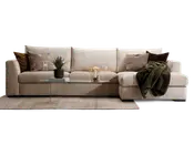 En moderne, lysebeige hjørnesofa med brede armlæn og bløde puder. Sofaen har en chaiselong på højre side og er pyntet med forskellige dekorative puder i neutrale og jordnære farver, herunder geometriske og teksturerede designs. Et mørkegrønt, blødt tæppe er lagt over chaiselongen. Foran sofaen står et slankt, rektangulært sofabord af glas med metalben, hvor der er placeret en potteplante, bøger, snacks og glas. Hele opstillingen står på et lyst, tekstureret tæppe, hvilket skaber en hyggelig og indbydende stue.