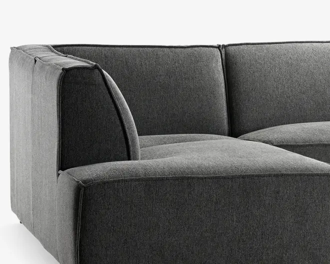 Mørkegrå polstret hjørnesektion med tykke, bløde sæde- og ryghynder. Stoffet er tæt vævet og giver et glat, moderne udtryk. Armlænet er bredt og polstret, hvilket matcher det firkantede og nutidige design. Syninger er synlige langs kanterne og tilføjer diskrete detaljer. Sektionen er designet til at kunne forbindes med andre modulære dele.