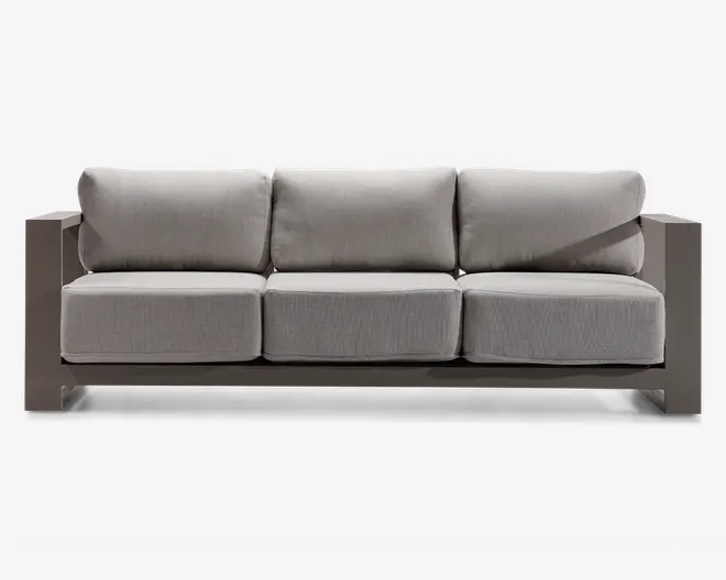 Moderne tresæders med tykke, lysegrå hynder på både sæde og ryg. Stellet er kantet og robust, lavet af et mat materiale i taupe eller lysebrun farve. Armlænene er brede og flade, hvilket giver ekstra overfladeplads. Designet er minimalistisk og nutidigt, velegnet til både indendørs og udendørs brug.