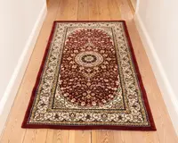 Rektangulært tæppe med en dyb rød baggrund og detaljerede blomster-mønstre i creme, guld og mørkeblå. Tæppet har et centralt medaljon-design omgivet af symmetriske motiver, og en bred cremefarvet kant dekoreret med snoede vinranker og blomsterdetaljer. Kanterne er afsluttet med en ensfarvet rød kant. Tæppet ser blødt ud og passer godt til en gang eller et smalt rum.