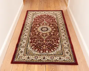 Rektangulært tæppe med en dyb rød baggrund og detaljerede blomster-mønstre i creme, guld og mørkeblå. Tæppet har et centralt medaljon-design omgivet af symmetriske motiver, og en bred cremefarvet kant dekoreret med snoede vinranker og blomsterdetaljer. Kanterne er afsluttet med en ensfarvet rød kant. Tæppet ser blødt ud og passer godt til en gang eller et smalt rum.