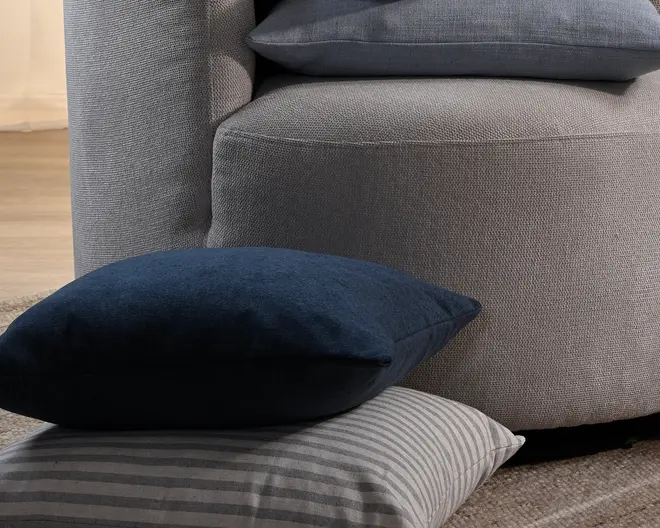 Nærbillede af en grå, tekstureret polstret sofa eller stol med en mørkeblå velourpude placeret oven på en gråstribet pude i forgrunden.