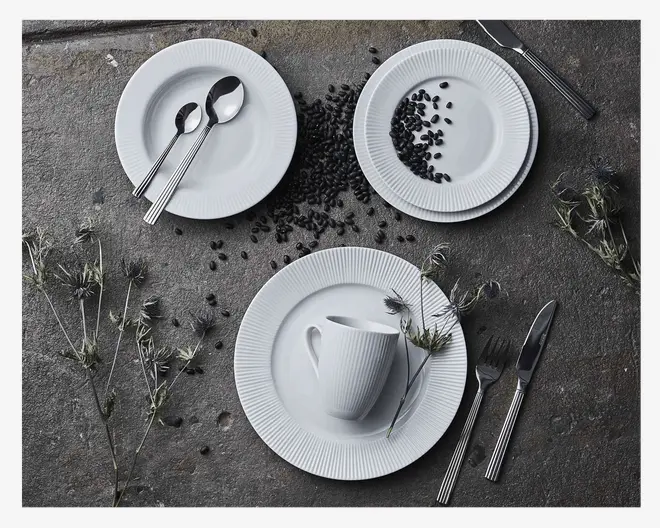 Billedet viser et sæt hvidt porcelænsservice med en diskret lodret rillet struktur. Sættet består af en middagstallerken, en mindre tallerken og en kop, alle med samme rillede design. Tallerkenerne og koppen er arrangeret på en mørk stenoverflade, ledsaget af bestik i rustfrit stål med et enkelt, moderne udtryk. Nogle tallerkener har kaffebønner spredt på sig, og der er tørrede blomsterstilke placeret omkring stellet, hvilket giver et rustikt præg til præsentationen.