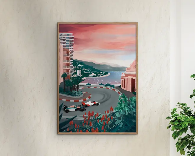 Indrammet kunstværk med en stiliseret illustration af et kystbylandskab ved solnedgang, hvor to Formel 1-racerbiler kører gennem et skarpt sving på en gadebane. Motivet viser moderne højhuse, frodige grønne planter med røde blomster i forgrunden samt udsigt over havet og fjerne bakker under en lyserød og orange himmel.