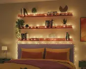 Tre træhylder på væggen over en seng er dekoreret med varme hvide lyskæder. Hylderne indeholder forskellige bøger, små potteplanter og dekorative genstande som en geometrisk skulptur og et moderne kunstobjekt. Sengen nedenunder har et lyse lilla polstret hovedgærde, sengetøj i sennepsgul og rustfarvede pyntepuder. Udtrykket er hyggeligt og indbydende med en blanding af moderne og naturlige elementer.