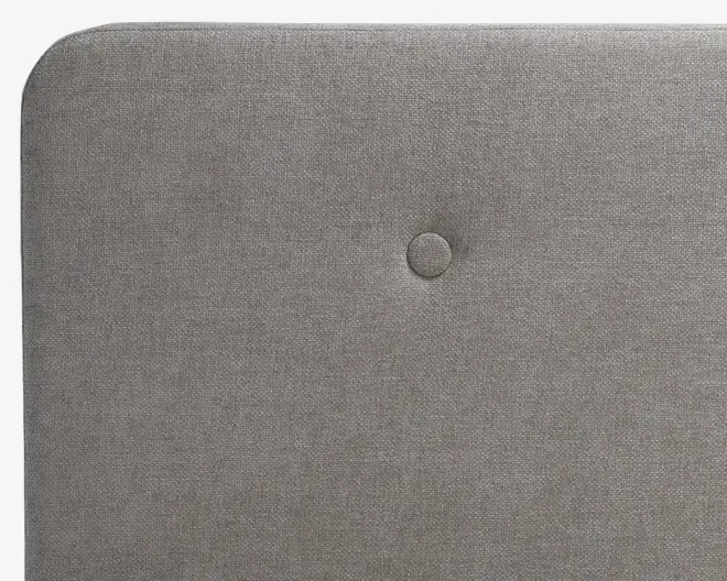 Nærbillede af en rektangulær, polstret overflade i lysegråt stof med en enkelt centralt placeret knap. Kanterne er blødt afrundede, og stoffet har en diskret vævet struktur.