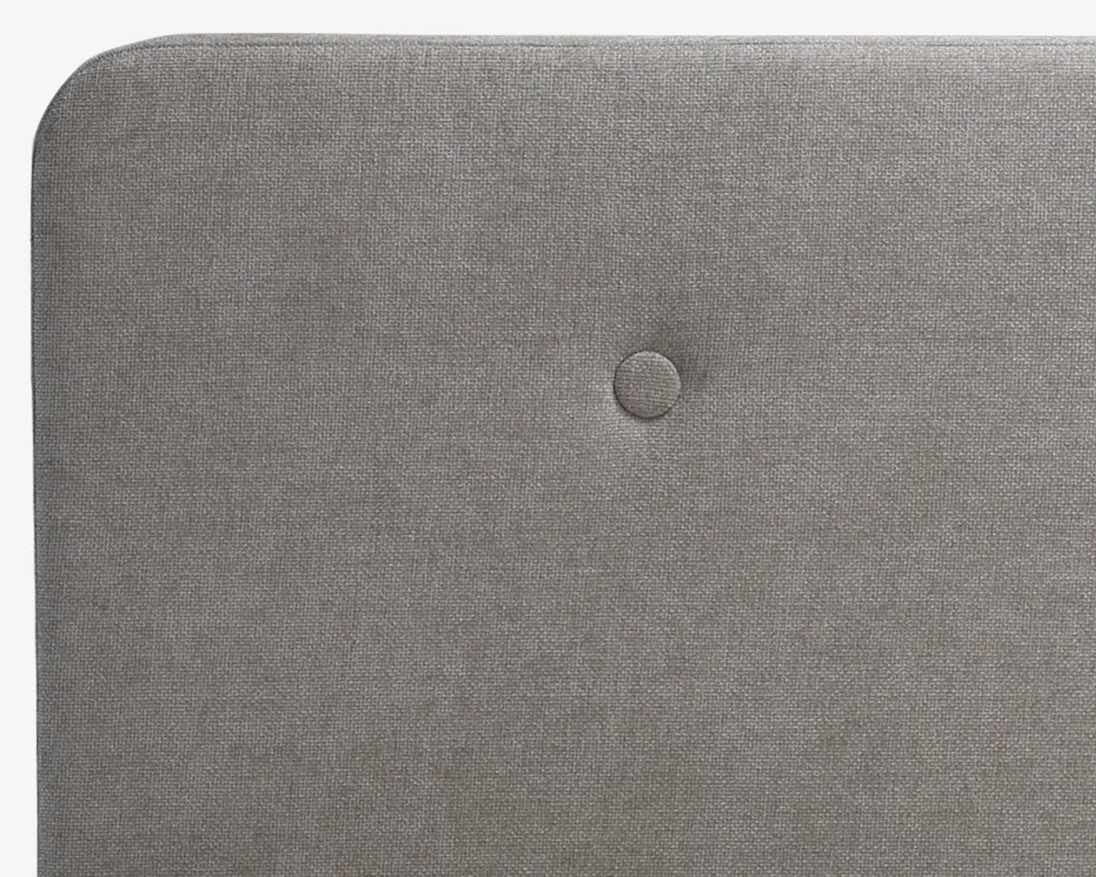 Nærbillede af en rektangulær, polstret overflade i lysegråt stof med en enkelt centralt placeret knap. Kanterne er blødt afrundede, og stoffet har en diskret vævet struktur.