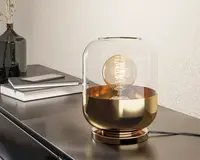 Moderne bordlampe med en rund glaskuppel, hvor den øverste halvdel er gennemsigtig og den nederste halvdel er i metallisk guld. Lampen har en synlig spiralformet glødepære indeni, hvilket giver et dekorativt udtryk. Foden er også i metallisk guld, og lampen står på en elegant, mørk overflade ved siden af stakke af bøger og en sort vase.