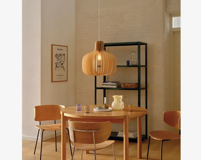 Et moderne spiseområde med et rundt bord i lyst træ med glat overflade, omgivet af fire matchende træstole med buede rygge og metalstøtter. Over bordet hænger en pendellampe med en skulpturel, lamelopbygget lampeskærm i træ, der spreder lyset varmt. På bordet står en hvid keramikkande, en matchende kop, et glas og et lille fad. I baggrunden ses en sort metalskænk med glas og bøger, og på væggen hænger et minimalistisk kunsttryk. Stilen er skandinavisk inspireret med lyse trætoner og rene linjer.