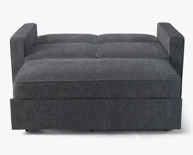 Et moderne møbel i mørkegråt stof med en firkantet form og rene linjer. Overfladen har polstrede detaljer, der danner fire store, bløde sektioner. Siderne har korte, firkantede armlæn, og stoffet ser ud til at være blødt og tekstureret. Designet er minimalistisk og funktionelt, velegnet til moderne indretning.
