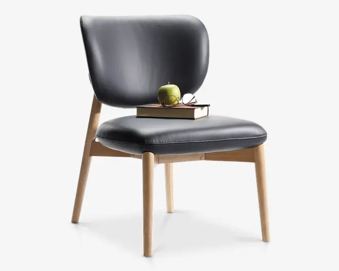 Stol med buet, polstret ryglæn og sæde betrukket med sort læder. Stel og ben er lavet af lyst træ med en naturlig finish, hvilket giver et moderne og minimalistisk udtryk. På sædet ligger to stablede bøger, et grønt æble og et par briller, hvilket indikerer komfort og funktionalitet.