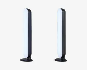 Et par slanke, lodrette LED-lysstænger med afrundede kanter, monteret på cirkulære sorte baser. Lysene har et moderne, minimalistisk design med en hvid oplyst front og en mat sort bagside. Velegnet til placering på skrivebord eller hylde og giver stemnings- eller accentbelysning.