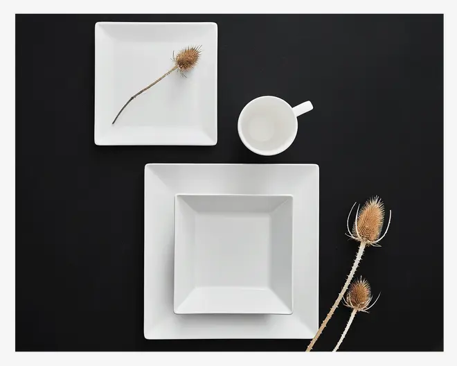 Et sæt moderne, hvide, firkantede tallerkener med rene, skarpe kanter. Sættet består af en stor firkantet tallerken, en mindre firkantet tallerken, en firkantet skål med skrå sider og et hvidt krus med et enkelt håndtag. Overfladerne er glatte og blanke, hvilket giver et minimalistisk og elegant udtryk. Produkterne er arrangeret på en sort baggrund, som fremhæver deres klare hvide farve.