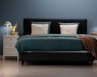 En moderne seng med en høj, polstret sengegavl i mørkt stof, udstyret med to store, teksturerede hvide puder og teal-farvede pudebetræk. Sengetøjet består af et teal dynebetræk og matchende lagner, med et brunt tæppe lagt over hjørnet. Sengen står på korte, mørke træben. Til venstre står et hvidt natbord med tre skuffer, en moderne lampe, en lille vase med gule blomster og en kop. Rummet har en minimalistisk stil med blå væg og lyst trægulv.