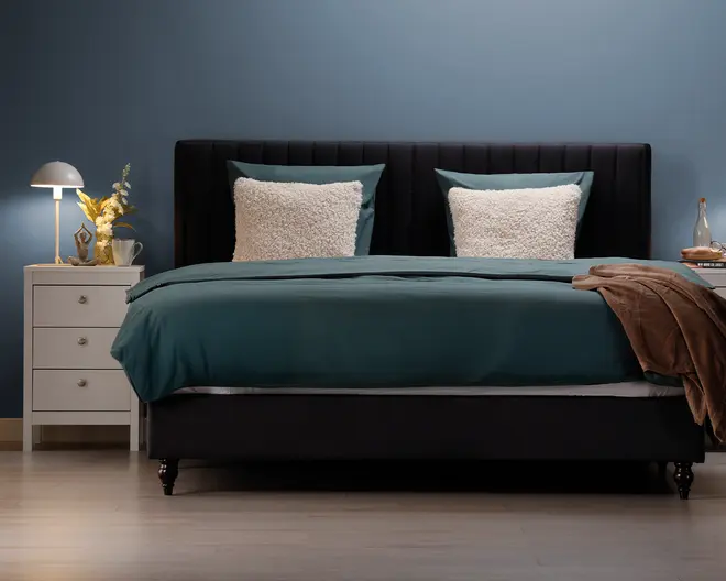 En moderne seng med en høj, polstret sengegavl i mørkt stof, udstyret med to store, teksturerede hvide puder og teal-farvede pudebetræk. Sengetøjet består af et teal dynebetræk og matchende lagner, med et brunt tæppe lagt over hjørnet. Sengen står på korte, mørke træben. Til venstre står et hvidt natbord med tre skuffer, en moderne lampe, en lille vase med gule blomster og en kop. Rummet har en minimalistisk stil med blå væg og lyst trægulv.