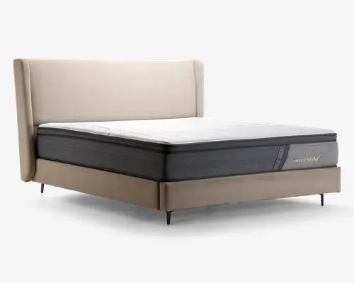 Sengen har et moderne design med en høj, polstret sengegavl i lysebeige stof. Madrassen er tyk og komfortabel med tofarvet udseende: det øverste lag er hvidt, mens den nederste del er mørkegrå med et diskret mærkelogo på siden. Sengerammen er betrukket med matchende beige stof og står på slanke, mørke ben, hvilket giver et elegant og moderne udtryk.