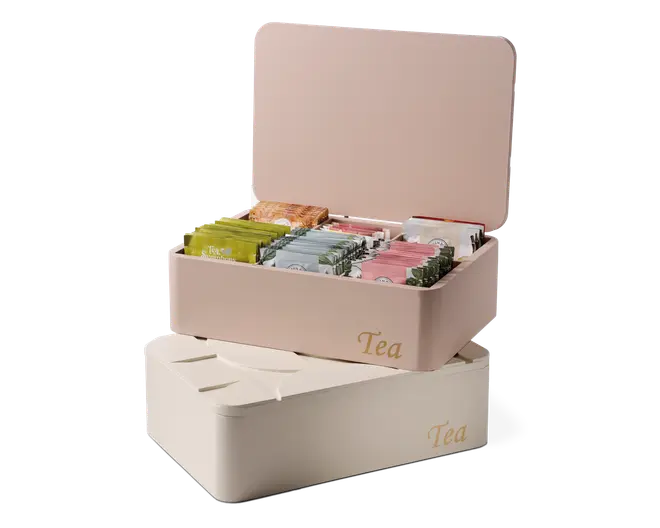 Rektangulære opbevaringsbokse med hængslede låg, mærket 'Tea' med gylden skrift på fronten. Den øverste boks er åben og viser flere rum, der organiserer forskellige individuelt indpakkede tebreve i forskellige farver. Boksene har en glat, mat overflade og fås i bløde pastelfarver, én i lyserød og én i creme.
