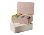 Rektangulære opbevaringsbokse med hængslede låg, mærket 'Tea' med gylden skrift på fronten. Den øverste boks er åben og viser flere rum, der organiserer forskellige individuelt indpakkede tebreve i forskellige farver. Boksene har en glat, mat overflade og fås i bløde pastelfarver, én i lyserød og én i creme.