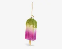 En dekorativ ornament formet som en ispind, med et farveforløb fra lysegul i toppen til kraftig pink i bunden. Toppen er dækket af grøn glimmer, der ligner drys eller hakkede nødder, og ornamentet hænger i en gylden snor. Overfladen er blank og glitrende, og en lille træpind stikker ud i bunden, som på en rigtig ispind.