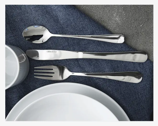 Et sæt blankt bestik i rustfrit stål bestående af en ske, en kniv og en gaffel. Hvert stykke har et enkelt, moderne design med glatte, afrundede håndtag og en skinnende, reflekterende overflade. Kniven har en diskret mærkeindgravering tæt på bladet. Bestikket er pænt arrangeret på et mørkeblåt tekstil ved siden af hvide keramik tallerkener og en kop.