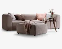 En moderne, L-formet sofa med afrundede kanter og blødt, lysebeige stofbetræk. Sofaen er pyntet med flere dekorative puder i nuancer af pink, brun og grå, hvilket tilføjer tekstur og komfort. Foran sofaen står en matchende puf med et hyggeligt, frynset tæppe lagt over. Ved siden af står et rundt sidebord i træ med en vase med blomster, stearinlys og en bakke med kopper og en skål, hvilket skaber en varm og indbydende stemning.