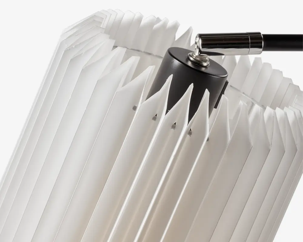 Nærbillede af en lampeskærm med geometrisk, plisseret design lavet af gennemsigtigt hvidt materiale. Skærmen har skarpe, kantede folder, der giver et tekstureret og moderne udtryk. Toppen af skærmen er fastgjort til en sort cylindrisk fatning med en metalarm, hvilket giver et nutidigt look.