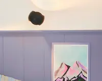 En moderne væglampe med mat sort finish er monteret over en væg, der er malet i en afdæmpet lavendelfarve. Under lampen hænger et indrammet maleri med stiliserede bjergtoppe i nuancer af pink, lilla og sort mod en lyseblå himmel. Maleriets ramme er lyse-lavendel og matcher vægpanelerne.