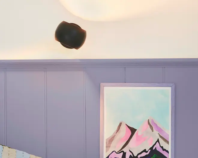 En moderne væglampe med mat sort finish er monteret over en væg, der er malet i en afdæmpet lavendelfarve. Under lampen hænger et indrammet maleri med stiliserede bjergtoppe i nuancer af pink, lilla og sort mod en lyseblå himmel. Maleriets ramme er lyse-lavendel og matcher vægpanelerne.