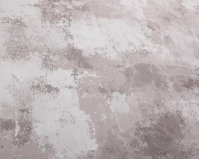 Billedet viser en nærbillede af en overflade med et abstrakt, tekstureret mønster. Designet har uregelmæssige pletter og penselstrøg-lignende områder i nuancer af beige, taupe og offwhite. Teksturen virker blød og let hævet, hvilket giver en følelse af dybde og dimension. Mønstret er ikke ensartet og skaber et moderne og kunstnerisk udtryk.