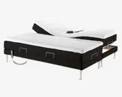 En moderne elevationsseng med to separate liggeflader, hver med en hvid, quiltet madras og sort bund. Sengen har metalben og metalbøjler ved fodenden af hver madras for at holde dem på plads. Den ene side af sengen er hævet ved hoved- og knæområdet for at vise justeringsmulighederne, og en fjernbetjening ligger på den hævede del.