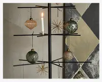 Nærbillede af en moderne, minimalistisk sort metaltræstruktur dekoreret med forskellige ornamenter. Ornamenterne omfatter grønne og gyldne glaskugler med indviklede mønstre, en rillet lyserød glasornament og gyldne stjerneformede dekorationer. Der er også et hvidt stearinlys, der holdes oprejst af en metalholder fastgjort til en af grenene. Baggrunden viser en væg med geometriske, malede mønstre i afdæmpede farver.