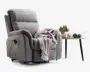 Blød, grå polstret lænestol med tyk polstring på sæde, ryg og armlæn. Stolen har en sidelomme med en synlig ledning, hvilket indikerer elektrisk recliner-funktion. Over armlænet og sædet ligger et tekstureret, frynset tæppe i bløde lilla og hvide nuancer. Ved siden af stolen står et lille træbord med sorte metalben, hvorpå der står en potteplante, et par briller, en kop og en skål.