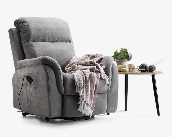Blød, grå polstret lænestol med tyk polstring på sæde, ryg og armlæn. Stolen har en sidelomme med en synlig ledning, hvilket indikerer elektrisk recliner-funktion. Over armlænet og sædet ligger et tekstureret, frynset tæppe i bløde lilla og hvide nuancer. Ved siden af stolen står et lille træbord med sorte metalben, hvorpå der står en potteplante, et par briller, en kop og en skål.