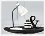 En hvid skrivebordslampe med blank overflade og fleksibel hals i krom, som gør det muligt at justere lysets retning. Lampen har en rund, solid fod og en konisk skærm. Den står på et sort fad sammen med en lille keramikvase med sorte og hvide vandrette striber samt et dekorativt sort ampersand-tegn.