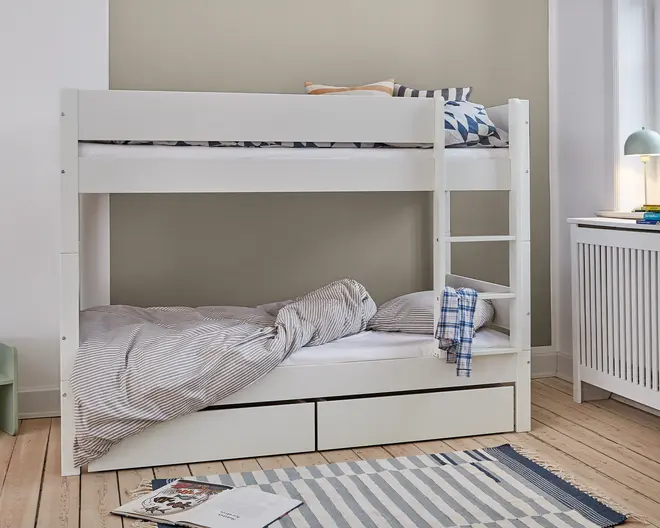 Hvid køjeseng i træ med et minimalistisk design, bestående af to enkeltsenge stablet oven på hinanden. Sengen har en indbygget stige i højre side for nem adgang til overkøjen. Begge køjer har enkle sengeheste for sikkerhed. Under den nederste køje er der to rummelige udtræksskuffer til ekstra opbevaring. Sengetøjet er i neutrale farver med stribede og geometrisk mønstrede puder, og et blåt ternet klæde hænger fra stigen. Sengen står på et lyst trægulv ved siden af et hvidt radiatorcover og en lille skrivebordslampe.