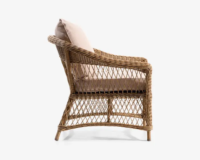 Sidebillede af en stol med stel i naturfarvet rattan og åbent, diamantformet fletmønster. Stolen har let tilbagelænet ryg og buede armlæn. Den er udstyret med tykke, lyse beige hynder på sæde og ryg for ekstra komfort. Benene er let udadbuede for stabilitet.