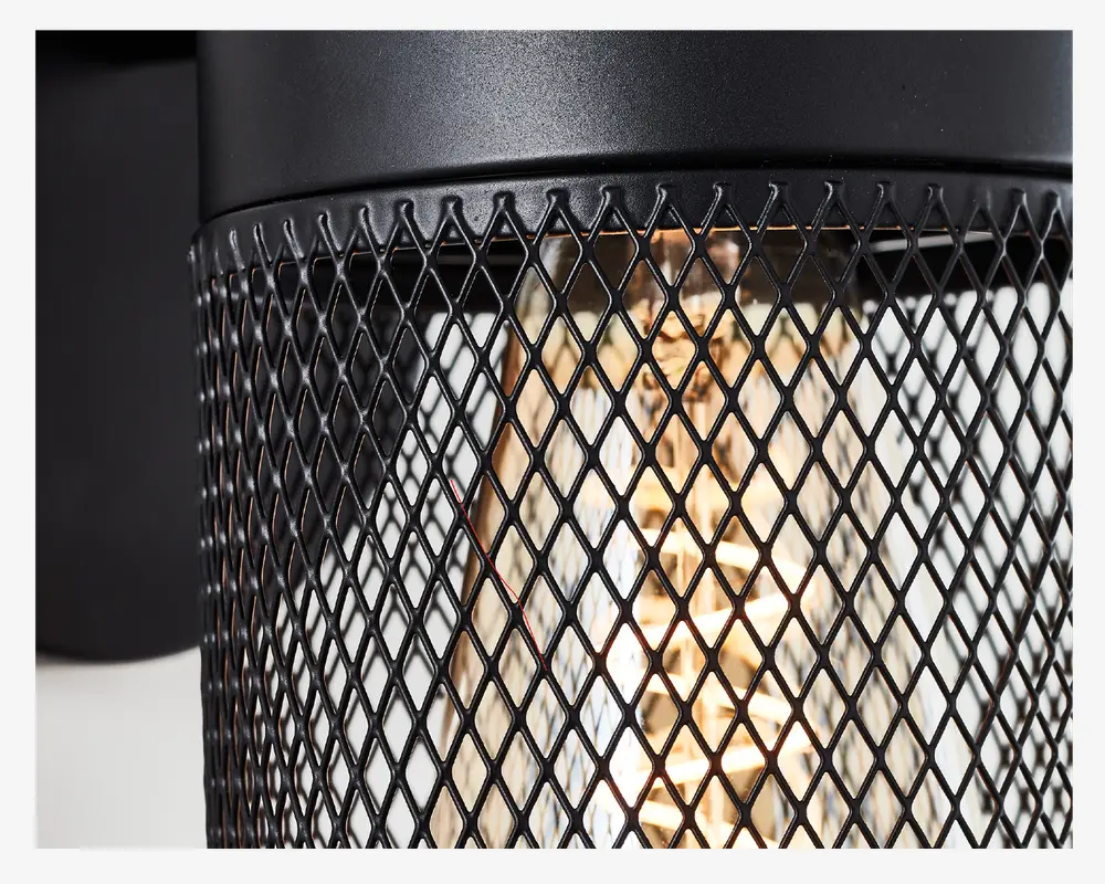 Nærbillede af en cylindrisk lampe med sort metalnet som skærm. Nettet har et diamantmønster, der lader det varme lys fra pæren indenfor skinne igennem. Lampen har en mat sort finish og et industrielt design.